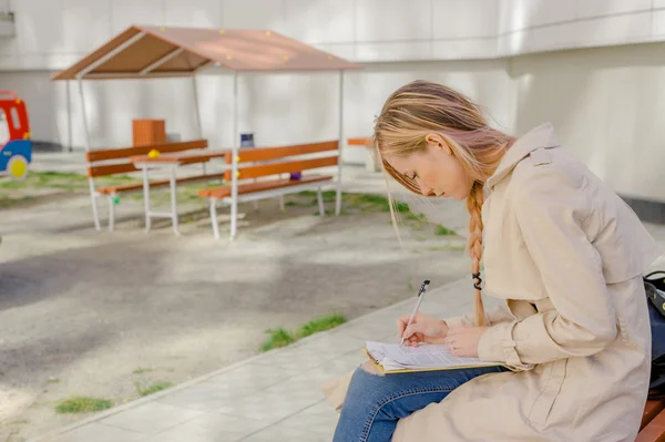 Young girl student draws and writes in a notebook sitting on a bench in the courtyard outside. combines work with study outdoor recreation. long hair, wearing a white T-shirt and blue jeans