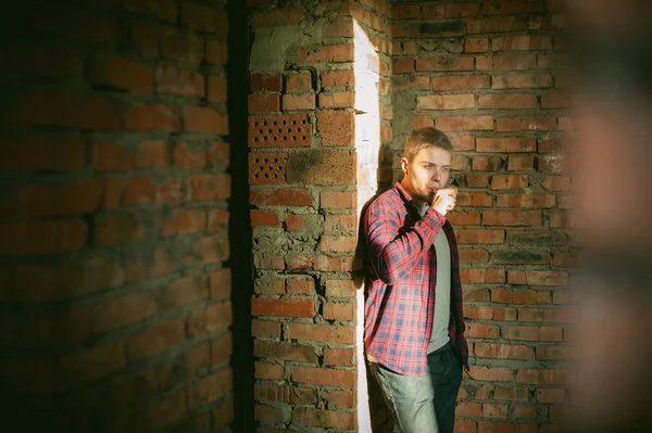 Man in a plaid shirt and jeans, smokes an electronic cigarette, blowing smoke and steam from the vaporization a mechanical device. location Abandoned unfinished building, brick walls, stylish.
