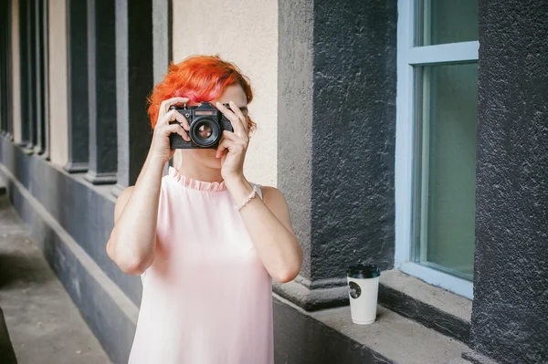 Portrait of a girl with a camera. woman with dyed red hair in a pale pink dress derzhet holding vintage camera