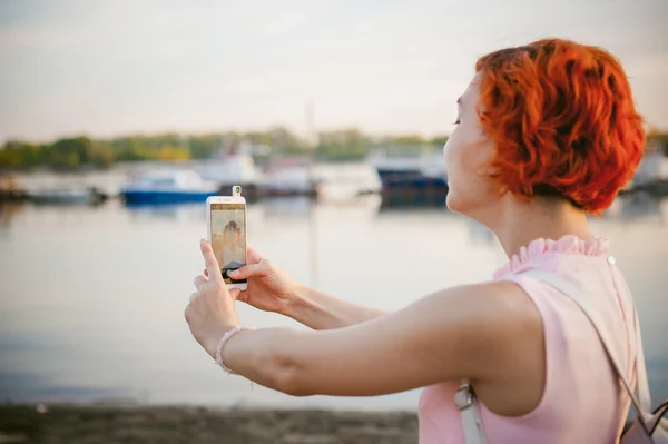 Girl in pale pink dress with red hair and backpack walking along river bank, pictures of themselves on their mobile camera phone, against backdrop of boats moored on a warm summer day