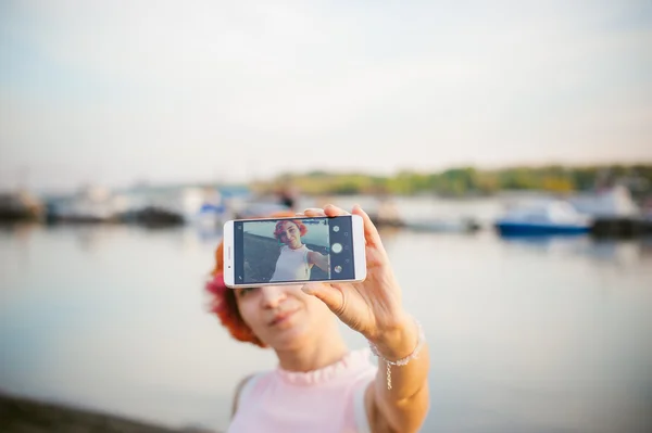 Girl in pale pink dress with red hair and backpack walking along river bank, pictures of themselves on their mobile camera phone, against backdrop of boats moored on a warm summer day