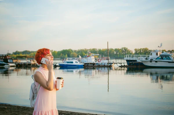 Girl in pale pink dress with red hair and backpack walking along river bank, talking on the phone and drinking coffee from a cardboard cup, against backdrop of boats moored on a warm summer day