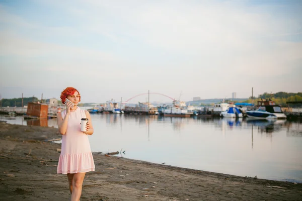 Girl in pale pink dress with red hair and backpack walking along river bank, talking on the phone and drinking coffee from a cardboard cup, against backdrop of boats moored on a warm summer day