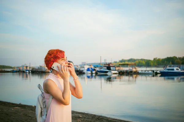 Girl in pale pink dress with red hair and backpack walking along river bank, talking on the phone and drinking coffee from a cardboard cup, against backdrop of boats moored on a warm summer day