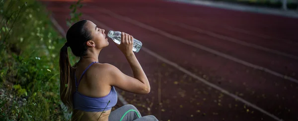 Young sporty woman drinking water after running in the stadium, close-up