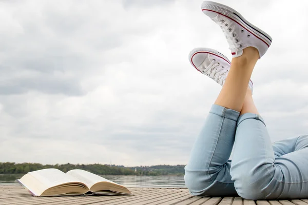 Girl lying with his legs crossed on a wooden pier next to a book