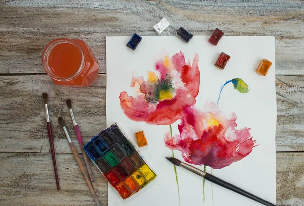 Watercolor paints, brushes and painted flowers on wooden background.