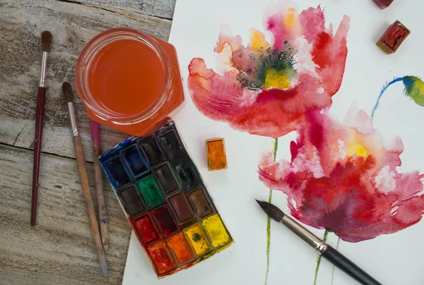 Watercolor paints, brushes and painted flowers on wooden background.