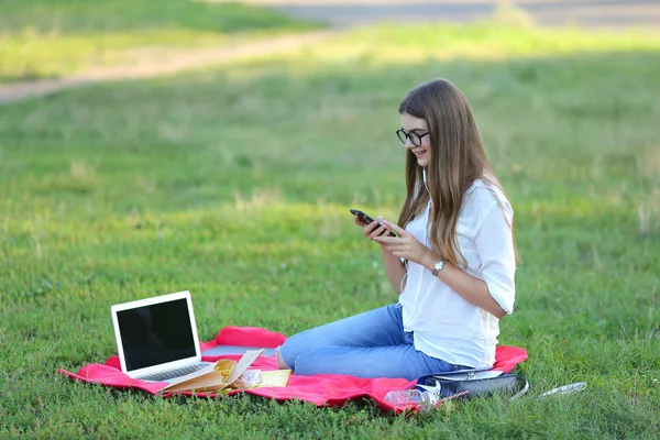 Young girl sitting in the park, smiling and working at his laptop,  listening to music