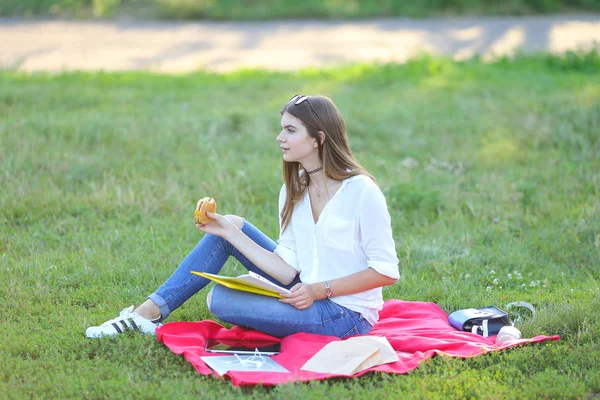 Young girl sitting on grass in the park and works at a laptop eating fast food
