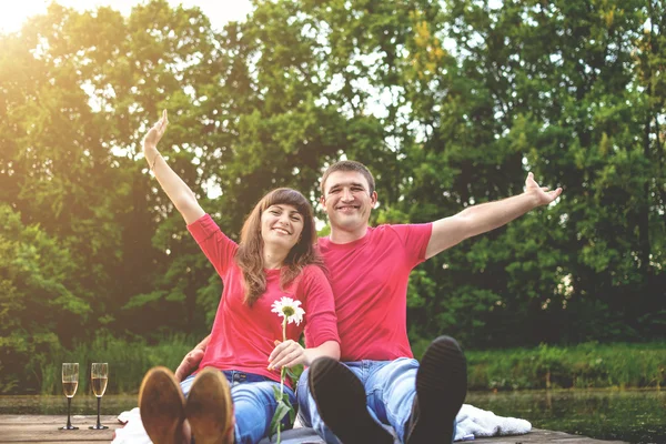 Cheerful pair young people in red shirts siyat by the river on a wooden bridge. A girl holding a white daisy. Toning