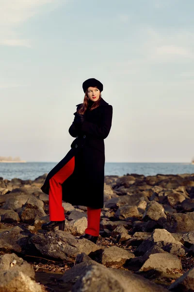 Girl in a black coat, in red pants, stands on large stones near the river in anticipation of Prince.