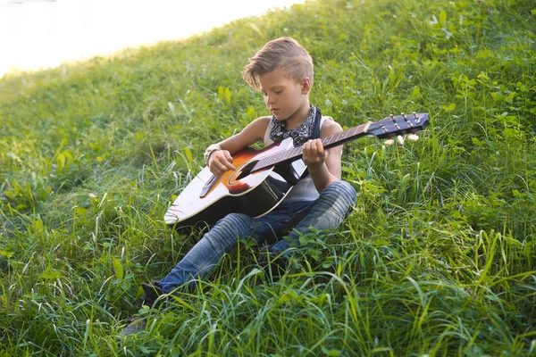 Little boy sitting on the grass and plays the guitar . A boy  learns to play the guitar .