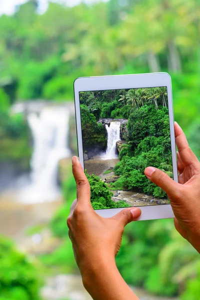 Female hand holding tablet and taking picture of scenic waterfall.