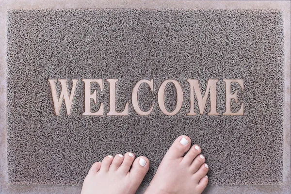 Welcome Door Mat With Female Feet. Friendly Grey Door Mat Closeup with Bare Woman Feet Standing. Welcome Carpet. Girl Feet with White Painted Toenails on Foot Scraper.