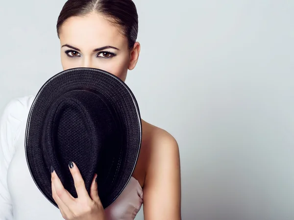Young dark-haired girl hiding her face behind black hat.
