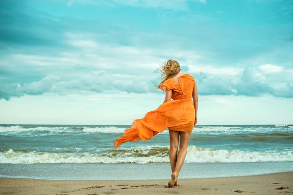 A young slender woman in orange dress is walking barefoot towards the storming sea