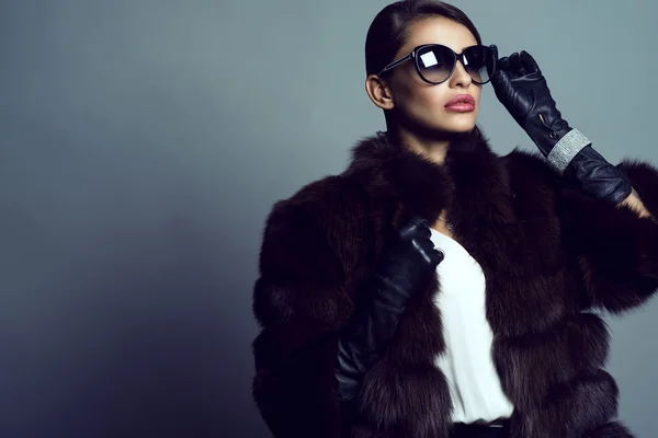 Beautiful glam model wearing white silk blouse, sable coat, leather gloves, sunglasses and set of luxurious bracelet, ring and silver chain with pendant.