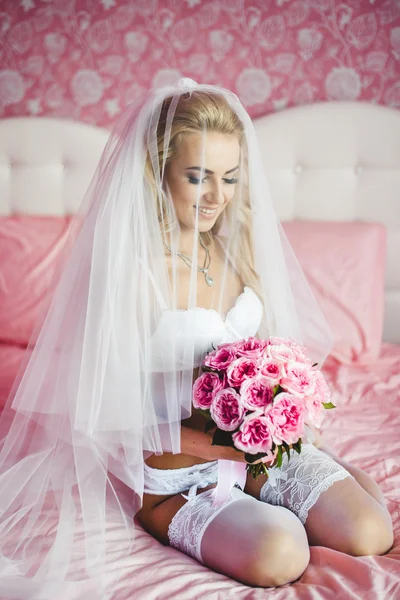 Beautiful sexy young bride underwear sitting on a bed with a bouquet of flowers in hands and with a veil on her head. Morning the bride.