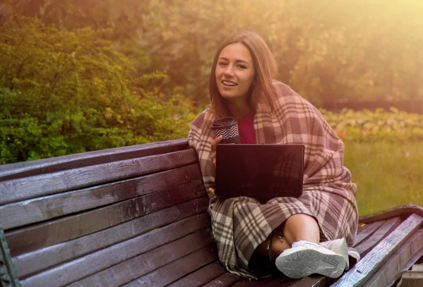 Charming girl wrapped in a blanket sitting on a bench in the park