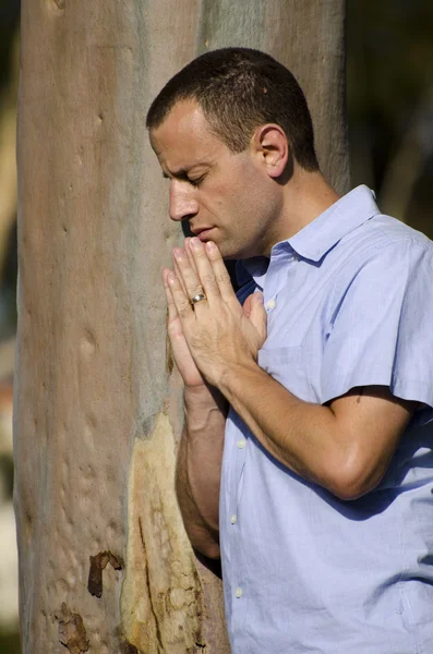 Man praying by a tree trunk outside in a park.