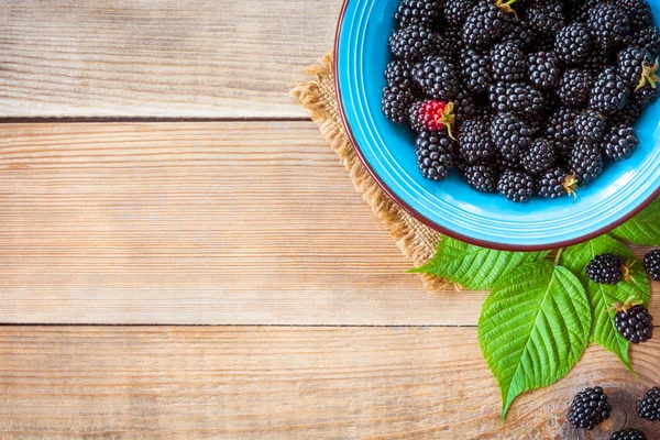 Fresh blackberries in blue ceramic bowl and leaves on wooden background in rustic style