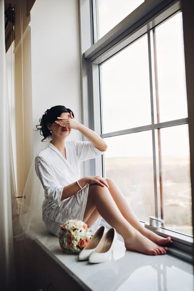 Woman by the window. Bride looking out the window, she waits for the groom. Beautiful bride in white wedding dress. elegant beautiful woman in white dress with creative hairstyle. shoes on the floor