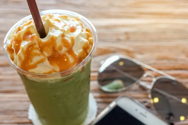 Cup of the matcha greentea frappe with caramel whipped cream on the brown bark beautiful texture background with warm light decorated with sunglasses and mobile phone