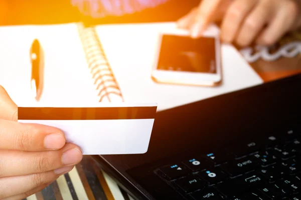 Online payment, man's hands holding a credit card over laptop and using smart phone on notebook for online shopping on the desk in the office, soft focus.