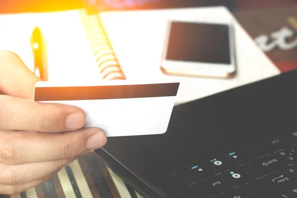 Online payment, man\'s hands holding a credit card over laptop and using smart phone on notebook for online shopping on the desk in the office, soft focus.