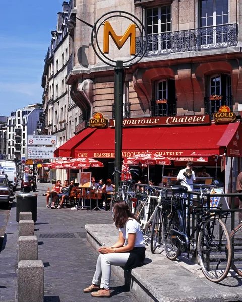 Young woman sitting by a Metro sign with a pavement cafe to the rear in the city centre, Paris.
