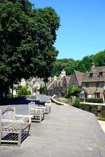 Stone cottages alongside the river Bybrook with wooden benches in the foreground, Castle Combe.