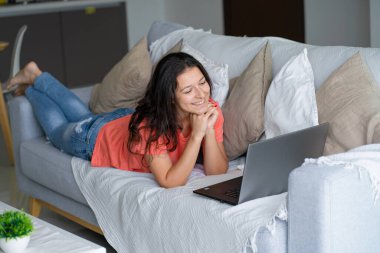 Kanepede uzanan kız dizüstü bilgisayara bakarken seviniyor. Gülümseme, iyi ruh hali, sevinç hissi..
