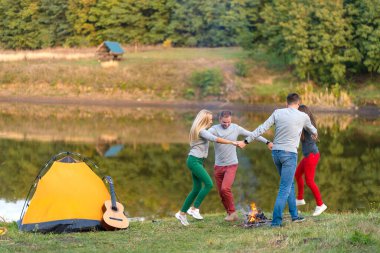 Gitarlı mutlu arkadaşlar, dışarıda eğleniyorlar, parkta göl kenarında dans edip zıplıyorlar güzel gökyüzü. Kamp eğlencesi.