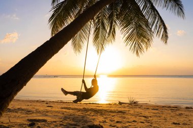 Adam ptropik plajda salıncakta sallanırken gün batımını seviyor. Salıncakta sallanan bir adamın siluetleri palmiye ağacına asılı, suda gün batımını seyrediyor.