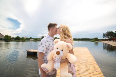 Romantik randevu sürprizi. Bir genç adam ve bir kız göle bakan bir iskelede. Adam kıza bir oyuncak ayı veriyor.