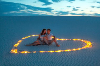 Romantic couple in love sitting on the sand in the desert. Evening, candles burn in the sand in form of heart around them. clipart
