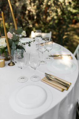 Pirinç bir vazoda taze çiçeklerle süslenmiş düğün masası. Düğün çiçekçisi olarak. Dışarıdaki yeşil manzaralı misafirler için bir ziyafet masası. Güllü buket, Eustoma ve okaliptüs yapraklı..