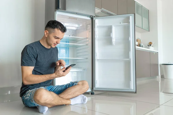 A young guy orders food using a smartphone. Empty refrigerator with no food. Food delivery service advertisement.