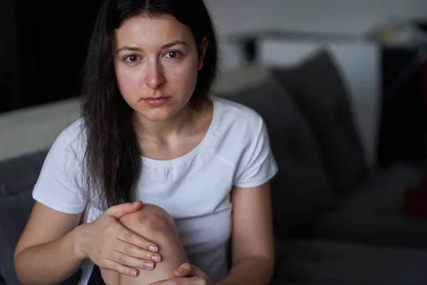 Portrait of beaten woman with tears of her face and bruise on knee sit on the sofa.