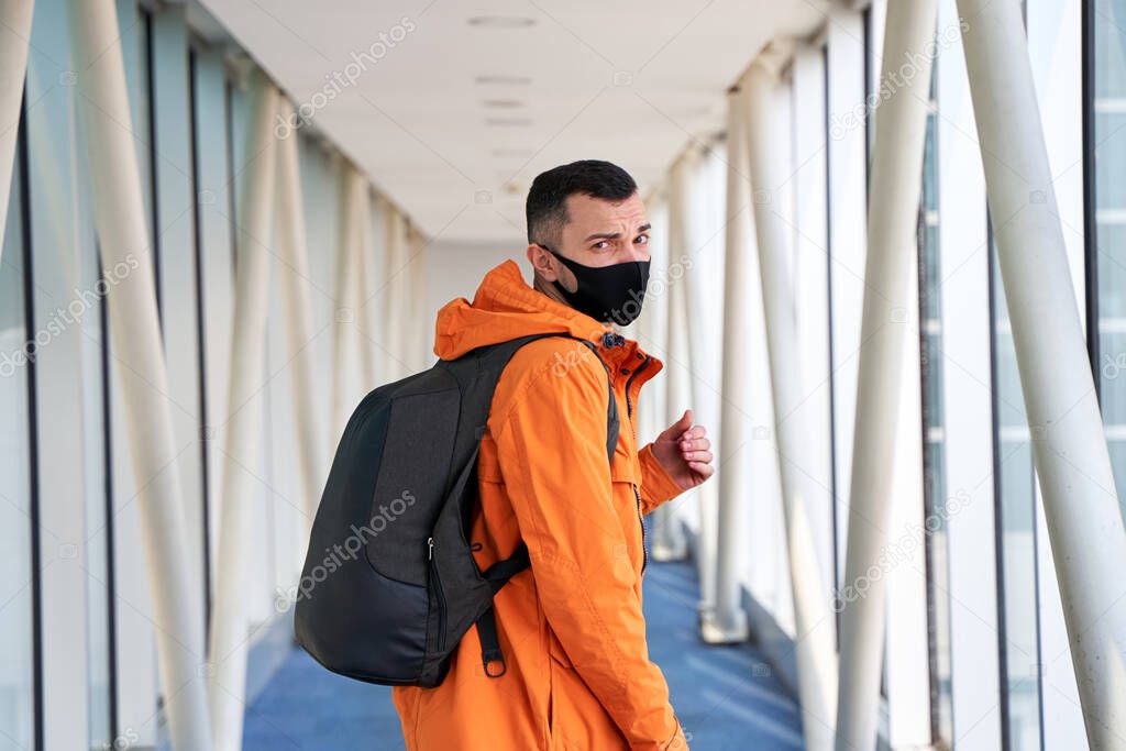 Guy tourist with a backpack and in a medical mask is walking along the airbridge from the plane to the airport terminal.