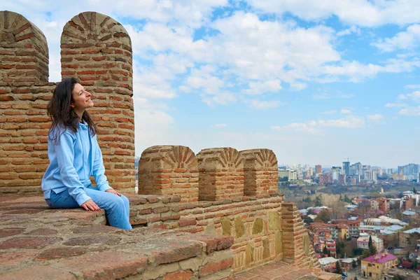 少女は街を見下ろす古代の要塞の壁に座っている間 景色と沈黙を楽しんでいます — ストック写真