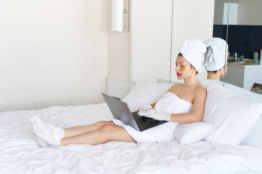 Bir duştan sonra, havluya sarılmış bir kız dizüstü bilgisayarda çalışır ve gözlerinin altındaki deri için kozmetik bantlar kullanır, ellerini ve ayaklarını nemlendirmek için dudaklar ve eldivenler. Evde vücut bakımı için kozmetik eğilimler.