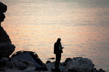 Yaz akşamları kamp çadırının yanındaki kayalık deniz kıyısında çay içen bir kız.