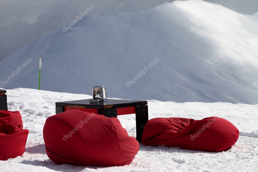 Red bean bag chairs and old table in outdoor cafe at ski resort and snow slope for freeriding at background. Caucasus Mountains in winter sun day, Georgia, region Gudauri.