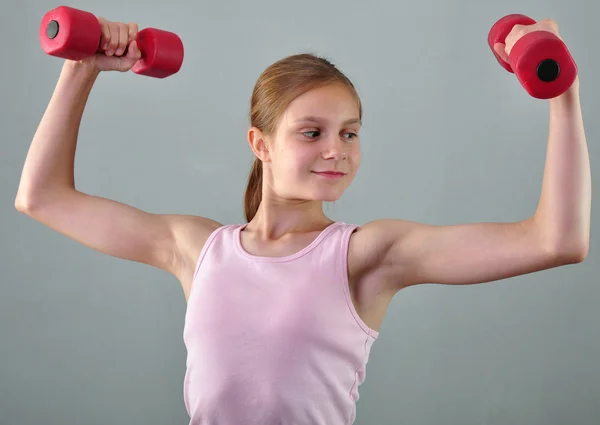 Adolescente menina esportiva está fazendo exercícios para desenvolver músculos em fundo cinza. Esporte conceito de estilo de vida saudável. Infância desportiva. Adolescente exercício com wieghts . — Fotografia de Stock