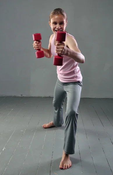 Chica deportiva adolescente está haciendo ejercicios con mancuernas para desarrollar con los músculos mancuernas sobre fondo gris. Concepto de estilo de vida saludable deportivo. Deportiva infancia. Adolescente haciendo ejercicio con pesas . — Foto de Stock