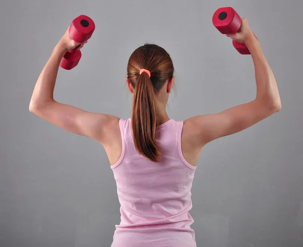 Teenage sportive girl is doing exercises to develop with dumbbells muscles on grey background. Sport healthy lifestyle concept. Sporty childhood. Teenager exercising with weights. — Stock Photo, Image