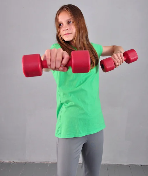Adolescente menina esportiva está fazendo exercícios com halteres para desenvolver músculos em fundo cinza. Esporte conceito de estilo de vida saudável. Infância desportiva. Adolescente exercício com pesos . — Fotografia de Stock