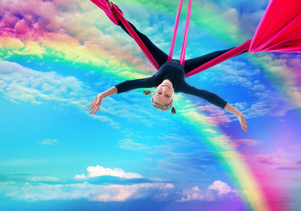 child hangs upside down on aerial silks in rainbow sky